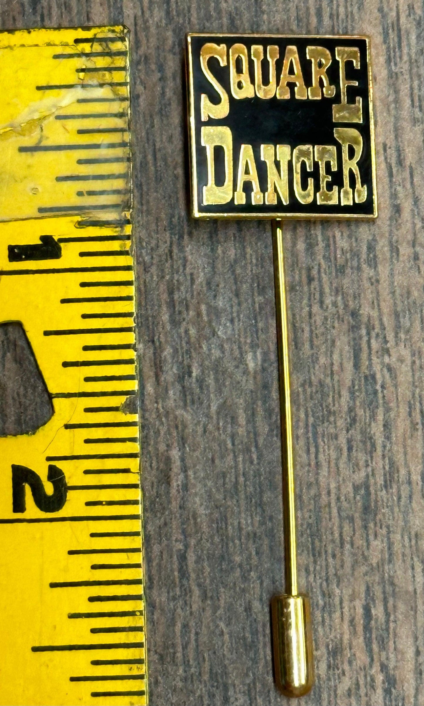 The Square Dancer Stick Pin by Square Up Fashions, a gold-colored pin reading "SQUARE DANCER," is shown beside a yellow tape measure indicating its 2-inch length, displayed on a wood surface—a stylish accessory for square dancers.