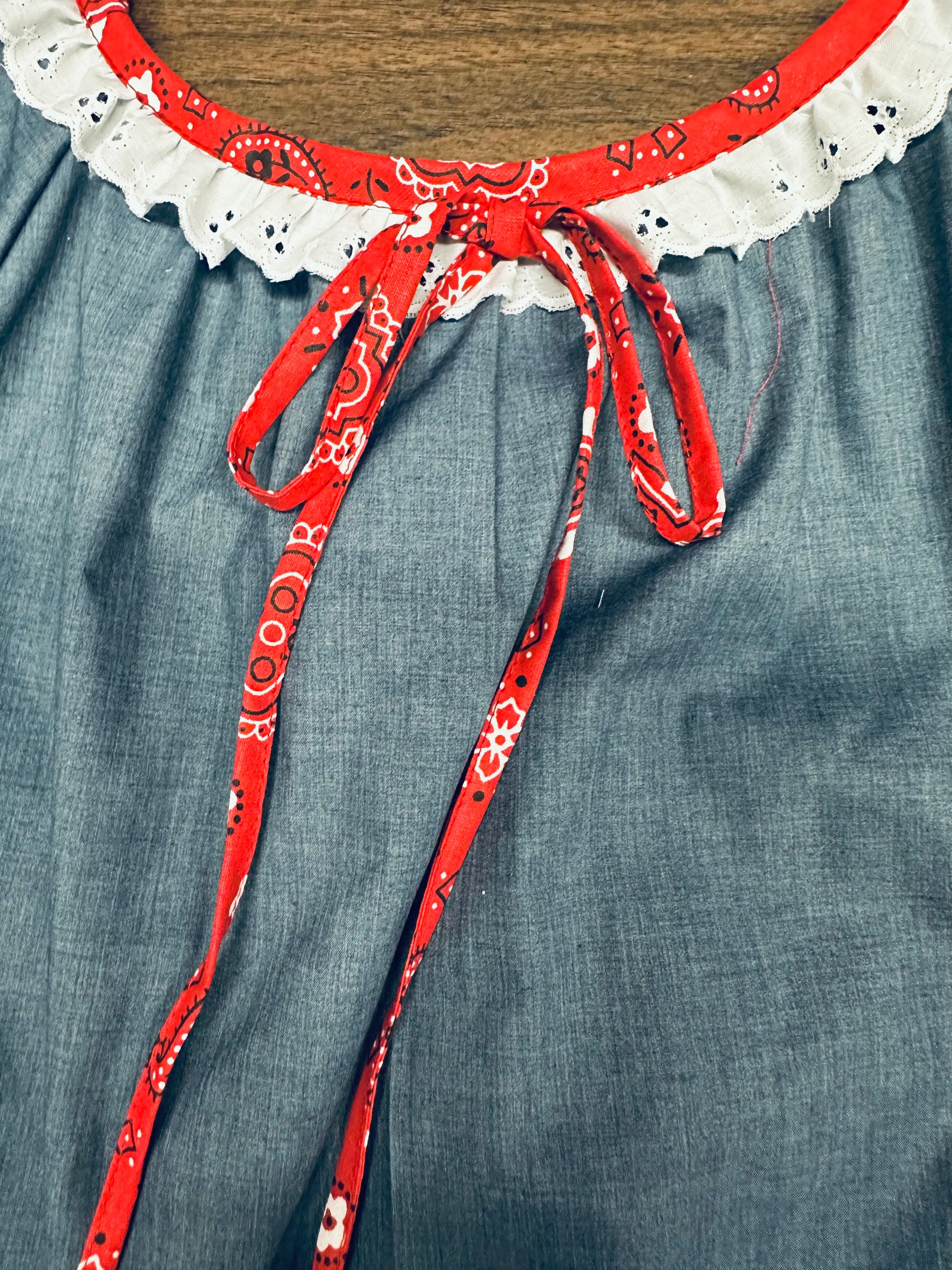Close-up of the Country Square Dance Outfit by Square Up Fashions, featuring a white eyelet lace neckline and red bandana print tie bow in classic peasant style, set against a blue fabric background.