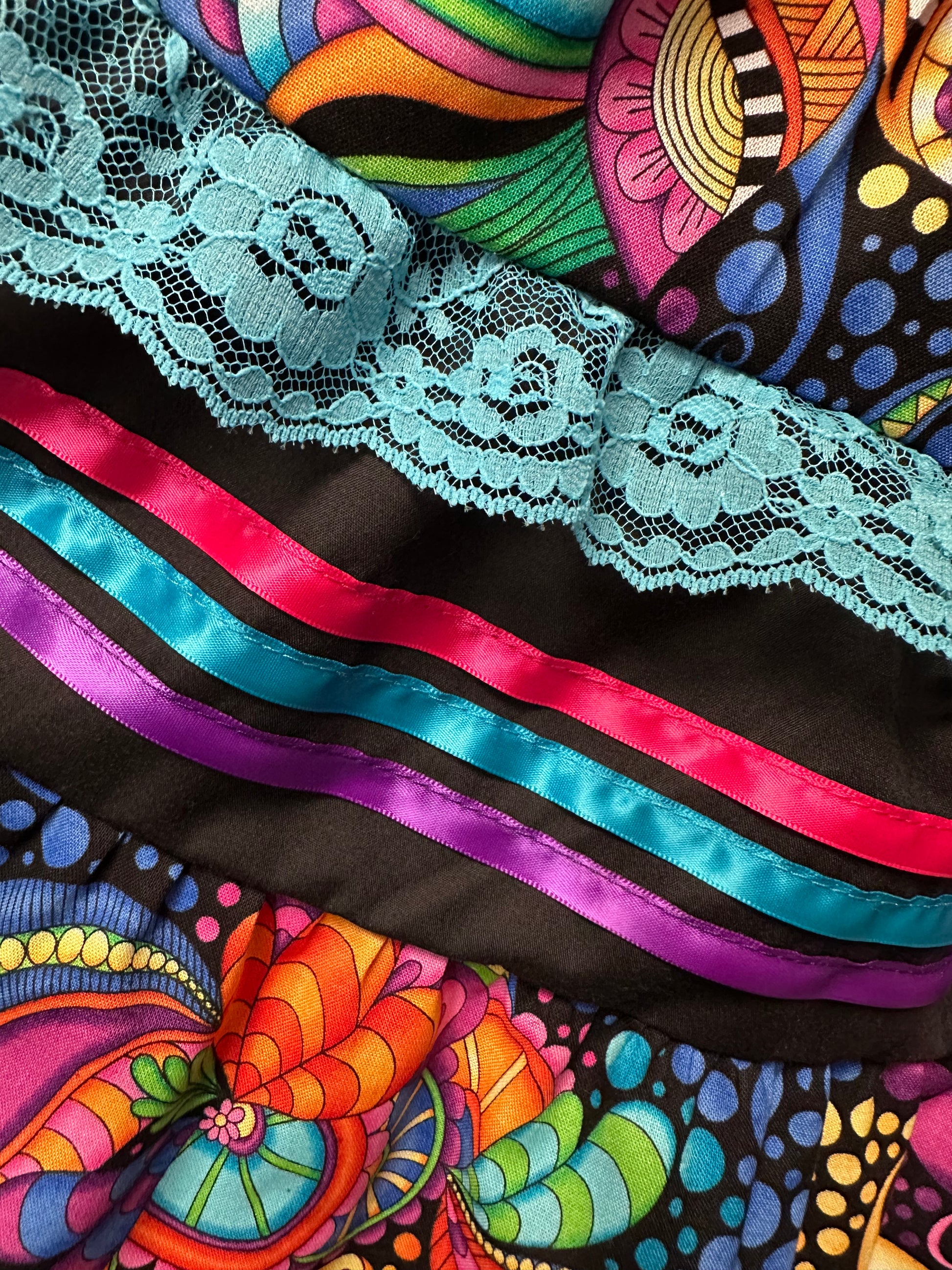 The Vivid Fantasia Square Dance Outfit by Square Up Fashions features bold abstract patterns in purple, orange, and green with blue lace trim and pink-blue satin ribbons—perfect for a lively skirt or festive blouse.