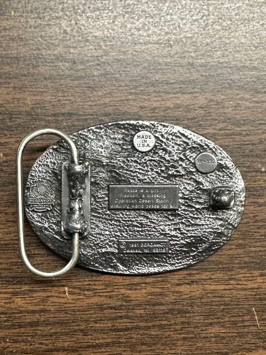 Back of a Desert Storm Square Dance Buckle by Square Up Fashions on a wooden surface, featuring multiple engravings, a metal loop, and an attachment hook.