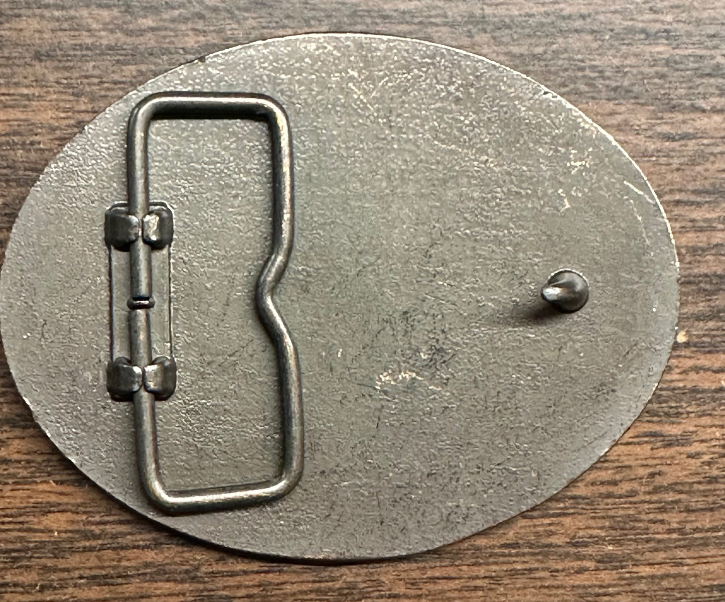 Back side of a Square Up Fashions Las Vegas Frontier Buckle, oval and metal with a loop and hook clasp, shown on a wooden surface.