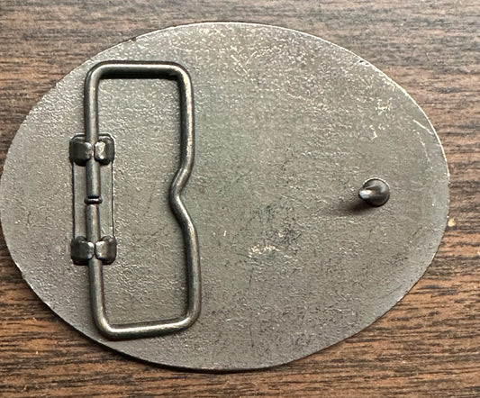 Back side of a Square Up Fashions Las Vegas Frontier Buckle, oval and metal with a loop and hook clasp, shown on a wooden surface.