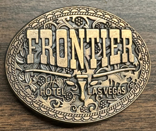 Las Vegas Frontier Buckle by Square Up Fashions features "FRONTIER HOTEL LAS VEGAS" in bold on oval brass with floral and scrollwork designs, displayed on a wooden surface.