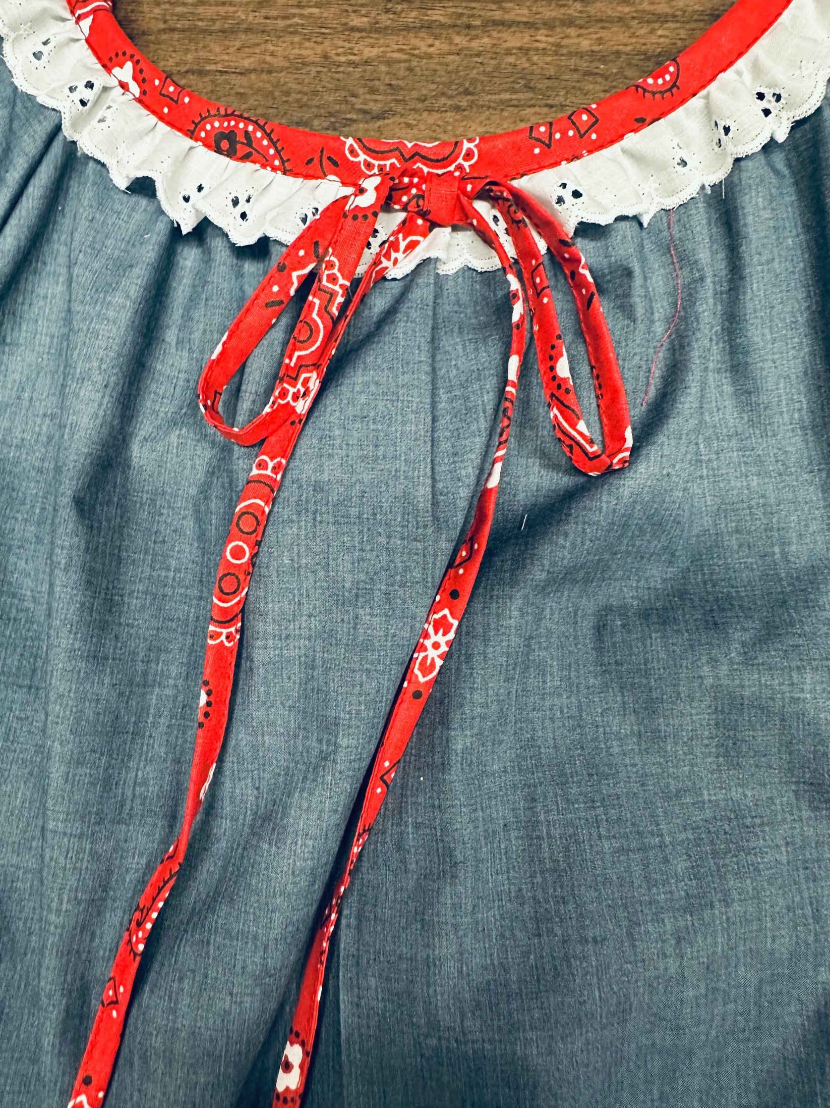 Close-up of the Country Square Dance Outfit by Square Up Fashions, featuring a white eyelet lace neckline and red bandana print tie bow in classic peasant style, set against a blue fabric background.