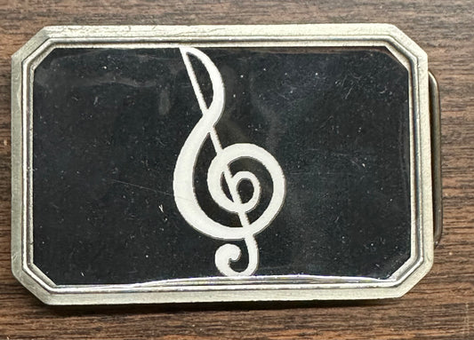 The Music Note Belt Buckle by Square Up Fashions is a rectangular metal accessory with a black background and a white treble clef, shown on a wooden surface.