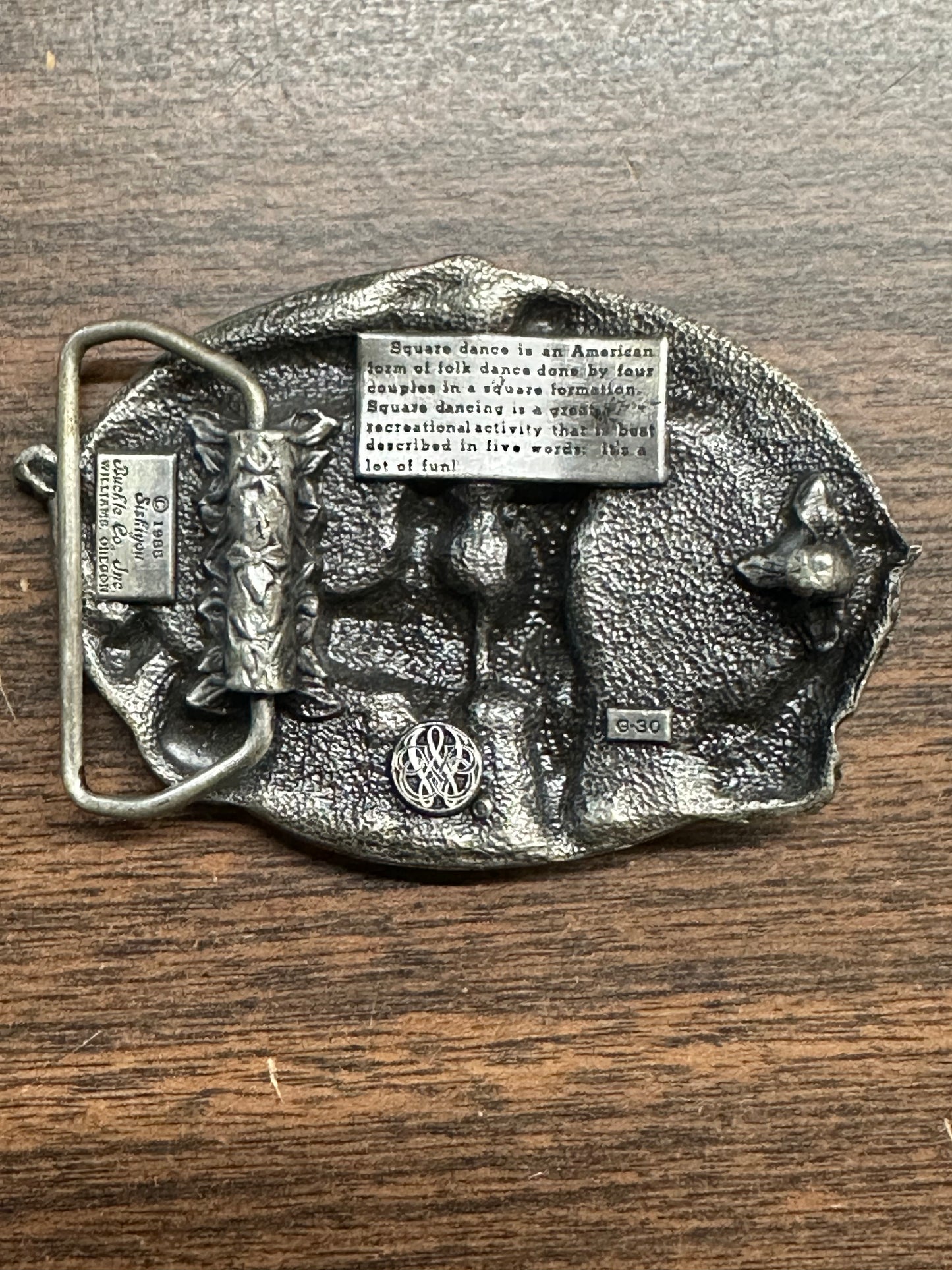 Square Up Fashions' Square Dance Buckle w/ instruments features an engraved definition, relief dancer, decorative patterns, and "DO SI DO." The buckle is shown resting on a wooden surface.