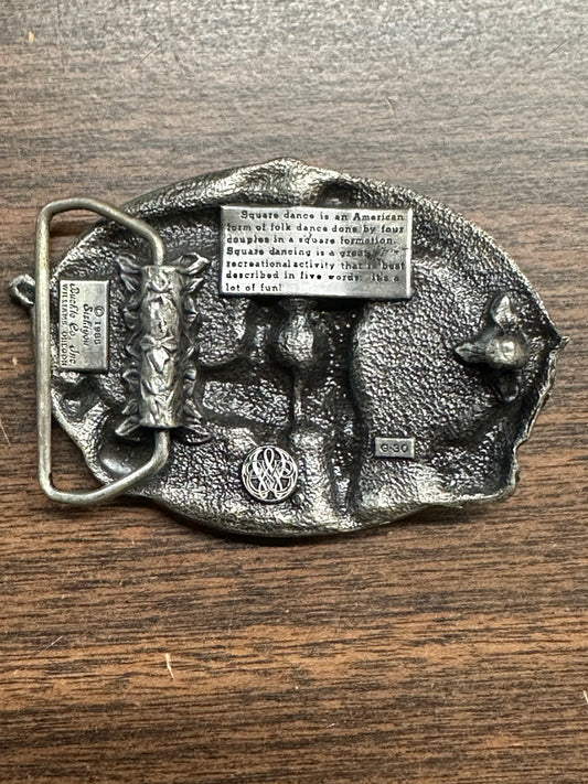 Square Up Fashions' Square Dance Buckle w/ instruments features an engraved definition, relief dancer, decorative patterns, and "DO SI DO." The buckle is shown resting on a wooden surface.