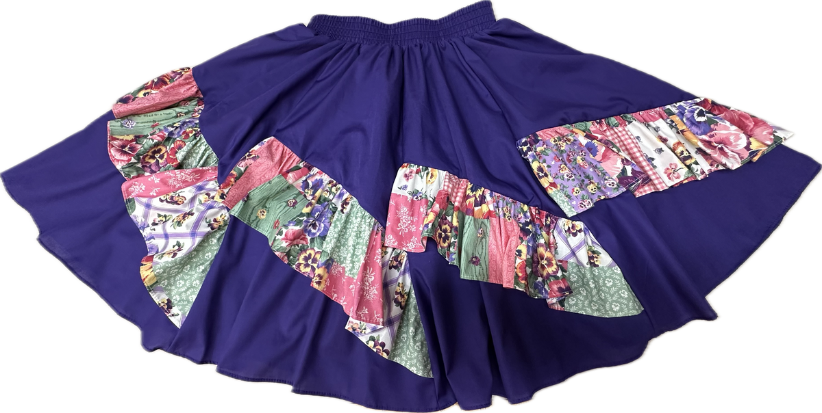 A colorful Diagonal Pinwheel Square Dance Skirt with vibrant print ruffle and floral designs displayed on a contrasting solid purple background by Square Up Fashions.