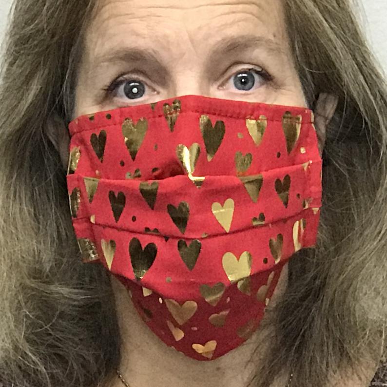 A woman wearing a Square Up Fashions red face mask with gold hearts on it. The face mask is washable and reusable, and features an adjustable nose wire for added comfort. It also provides 2 layers of