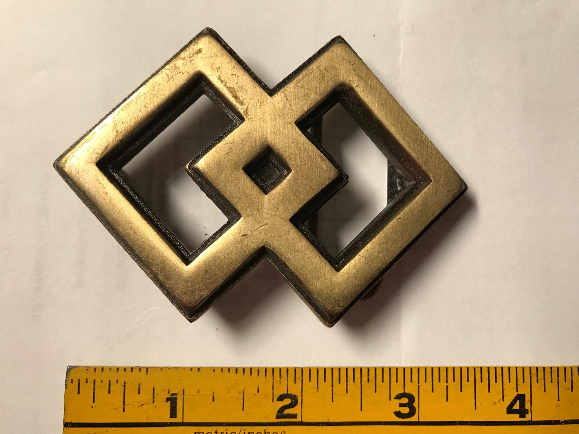 A Square Dance Buckle with an interlocking design, positioned next to a ruler, made by Square Up Fashions.